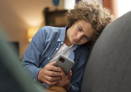 Jeune garçon a l'aire triste assis sur un canapé et regradant son téléphone