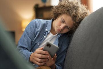 Jeune garçon a l'aire triste assis sur un canapé et regradant son téléphone